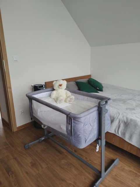 Gray bedside bassinet with cream teddy bear next to a wooden bed in a white bedroom with wooden floor