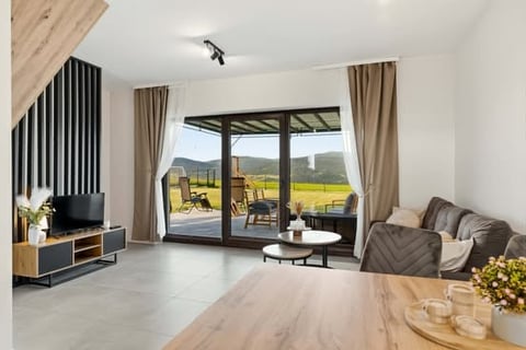 Modern living room with light wood flooring, gray sofa, mounted TV, floor-to-ceiling glass doors opening to patio with mountain views, neutral color scheme with wooden accents