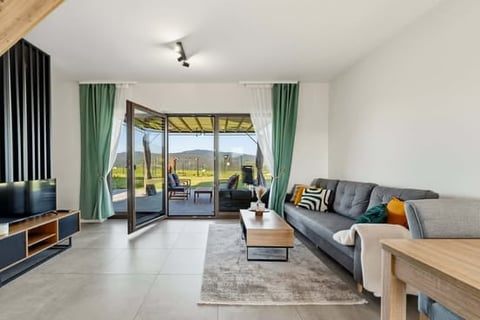 Modern living room with gray sofa, wooden furniture, large glass doors opening to patio, and mountain views
