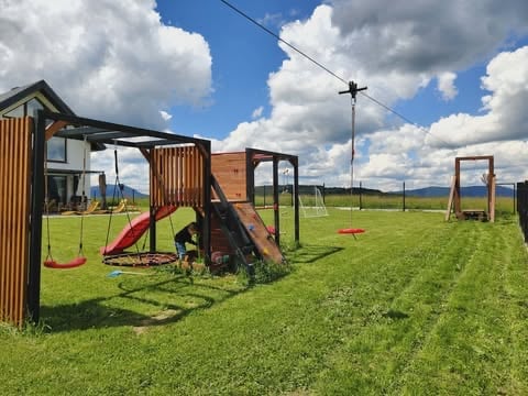 Outdoor playground equipment in a grassy field with mountains visible in the distance under a blue sky with white clouds
