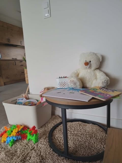 Childs play area with white teddy bear on round desk, colorful toy blocks, storage bin, and books on a beige area rug