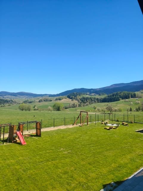 Rural playground with red slide and wooden structures on green grass overlooking rolling hills and forested mountains under clear blue sky