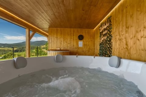 Modern wooden hot tub with mountain view, situated on a covered deck with towels and landscape scenery visible through large windows