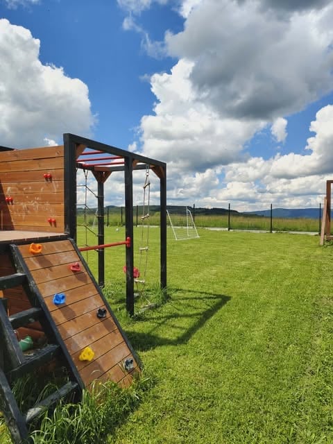 Outdoor playground equipment in a grassy field with blue sky and clouds, including a climbing wall and swing frame structure