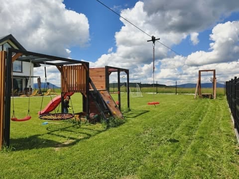 Outdoor playground with wooden structures, red slide, swings, and climbing frames on a green grass field under a blue sky