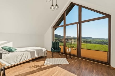 Modern bedroom with wood flooring, bed, wooden-framed windows overlooking green rolling hills and countryside landscape