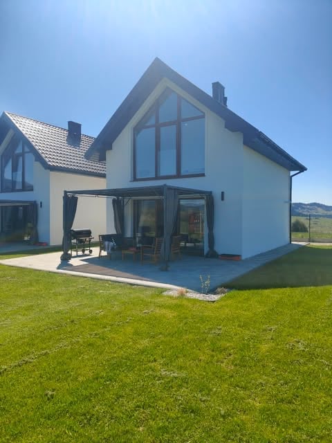 Modern white house with large gabled windows, blue roof trim, and patio overlooking green lawn and distant mountains under clear blue sky.