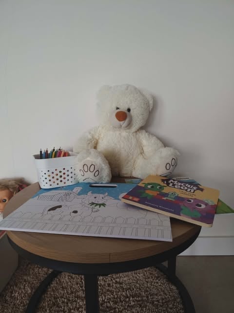 Cream-colored stuffed bear with red nose sitting on a table surrounded by coloring pages, colored pencils, and childrens books