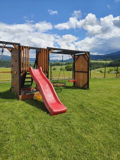 Wooden playground structure with red slide and swing set on green grass with mountain valley landscape in background