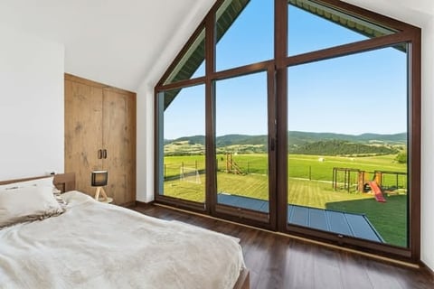 Modern bedroom with large multi-pane windows overlooking rolling green pastures and mountains, featuring a bed and wooden door