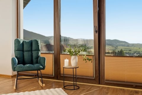 Green upholstered chair and side table on wooden balcony overlooking mountain landscape with valley and pine trees