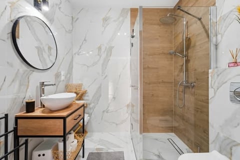 Modern bathroom with marble walls, wooden vanity with white bowl sink, circular mirror, glass shower enclosure, and toilet