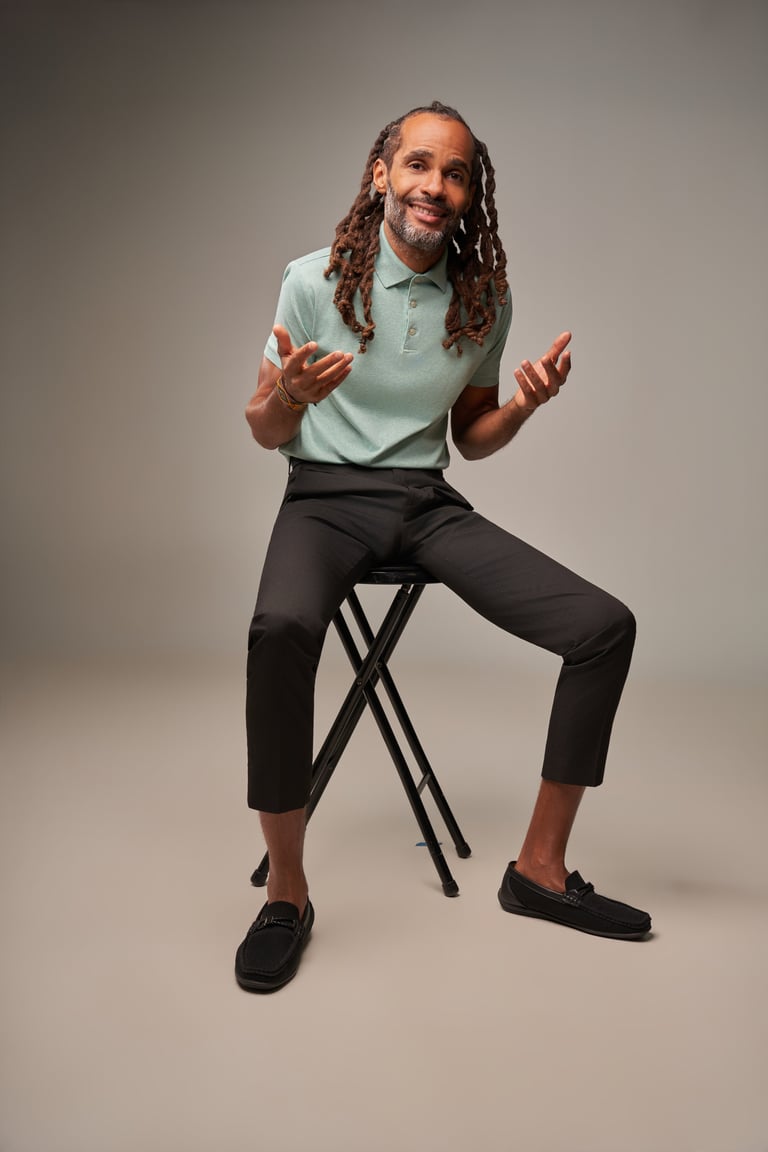 Man with long dreadlocks sitting on a stool, wearing a light blue polo shirt and black pants, gesturing with both hands and smiling at the camera