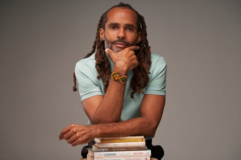 Man with dreadlocks wearing light blue shirt resting chin on hand, leaning on stack of books against gray background