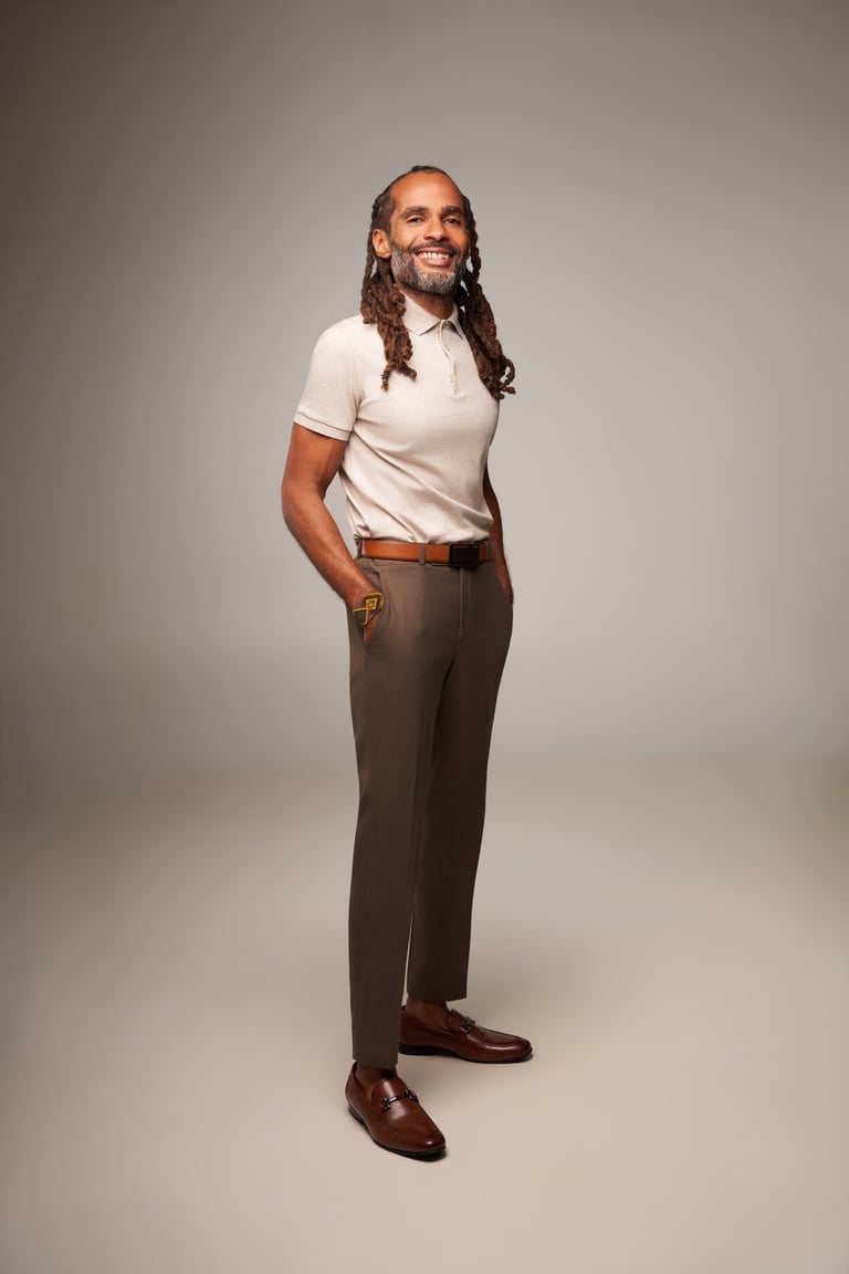 Man with long dreadlocks wearing white polo shirt, brown pants, and burgundy loafers, smiling at camera against gray background