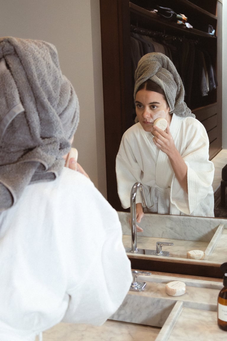 Woman in bathroom mirror wrapped in white bathrobe and towel, applying skincare product to face