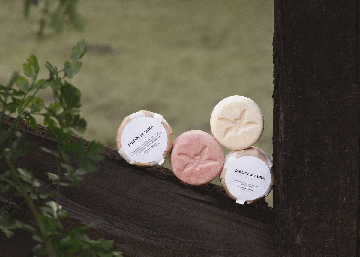 Four handmade soaps in white and pink arranged on a dark wooden ledge with green plant foliage on the left side