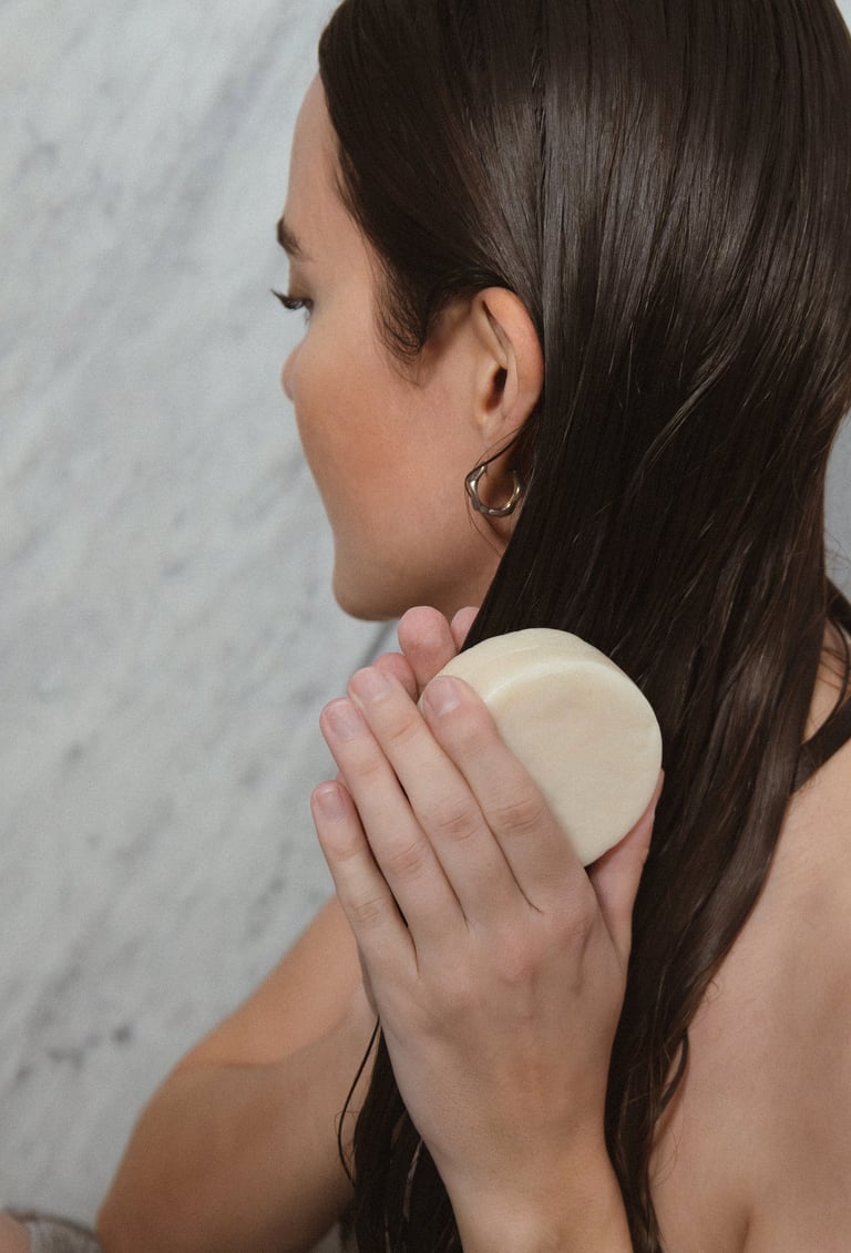 Woman with long dark hair holding a round cosmetic product against her shoulder, shown in profile view
