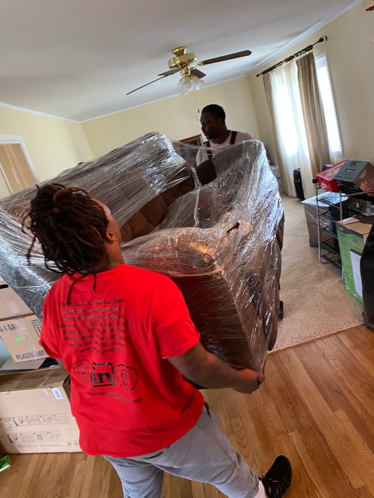 Two people wrapping a couch with plastic in a bedroom during furniture moving preparation