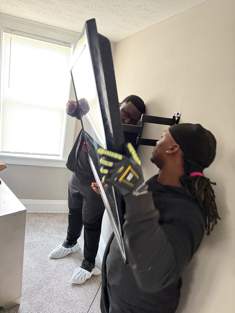 Two people installing a flat-screen TV on a wall in an indoor room with a window and white walls.