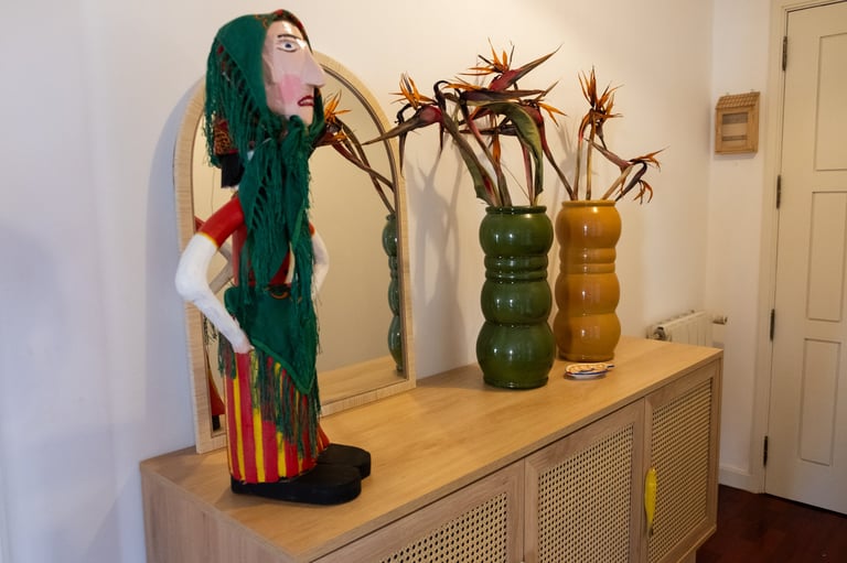 Decorative doll with teal hair and colorful dress stands beside vases with dried flowers on a wooden cabinet