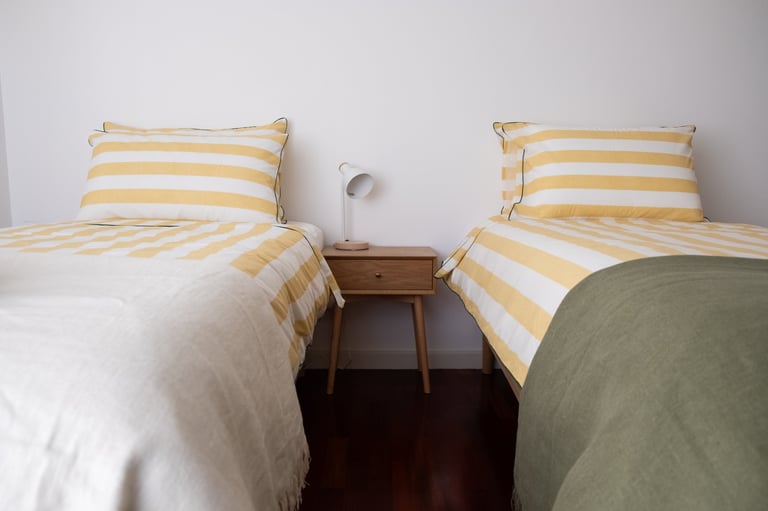 Two twin beds with yellow and white striped bedding separated by a wooden nightstand with a lamp in a minimalist bedroom