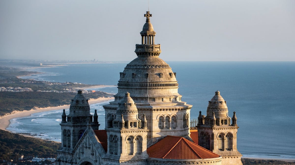 Aerial view of a grand basilica with golden dome and red roof overlooking a coastal landscape with blue ocean and beaches
