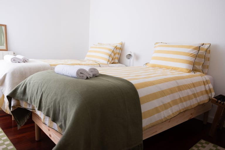 Two beds with yellow and white striped bedding and throw pillows in a bright, minimalist bedroom with white walls