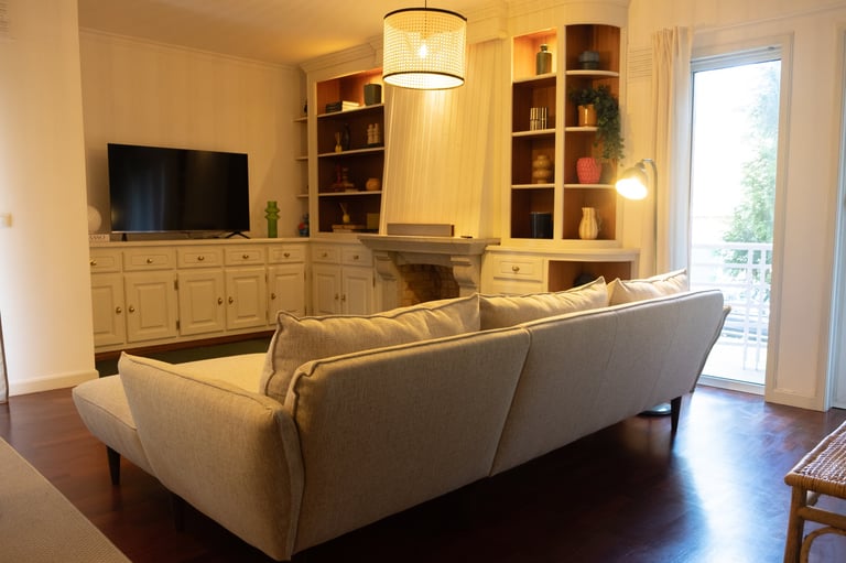 Modern living room with beige upholstered sofa, wooden built-in shelving, TV console, and glass doors opening to a patio
