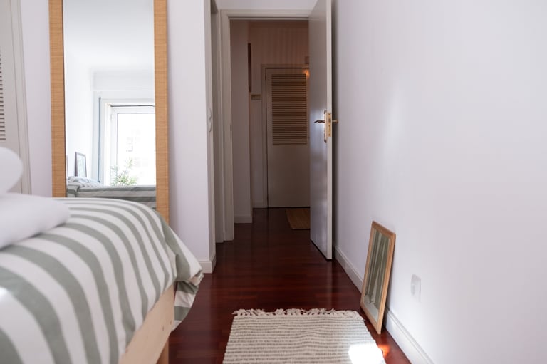Narrow bedroom with striped bedding, wooden door, dark hardwood floor, and a small rug against white walls