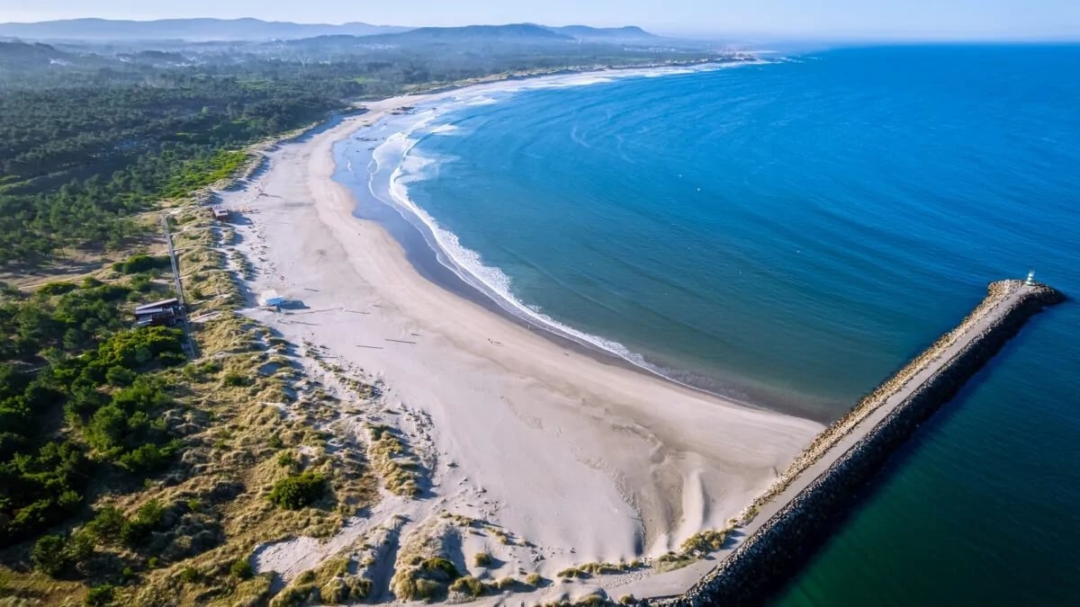 Aerial view of a sandy beach with a wooden pier extending into blue ocean waters, bordered by green forest and mountains