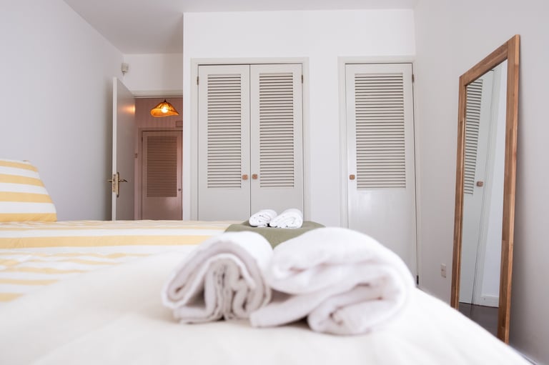 Clean hotel bedroom with rolled white towels on bed, louvered closet doors, and wood-framed mirror