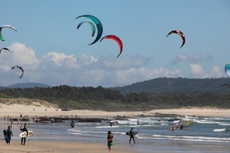 Multiple kitesurfers with colorful kites above a beach with people in the water and forested hills in the background
