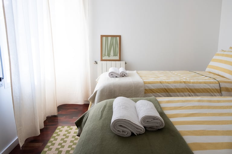 Bright, minimalist bedroom with two beds topped with striped bedding and rolled towels, sheer white curtains, and simple wall art