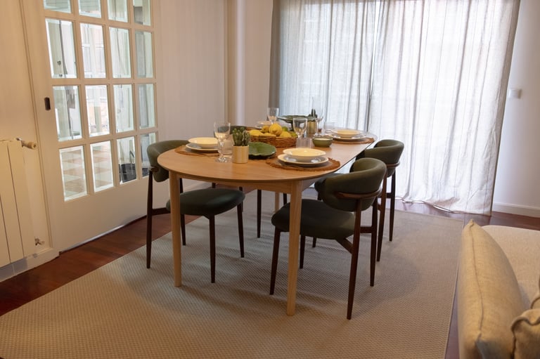 Modern dining room with oval wooden table set for two, black chairs, and floor-to-ceiling windows with sheer curtains
