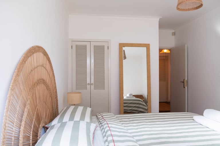 Modern bedroom with striped bedding, woven rattan headboard, wooden-framed mirror, white louvered closet doors, and pendant lighting