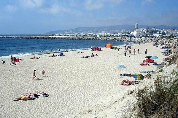 Crowded beach with sandy shore, beachgoers, colorful umbrellas and tents, coastal cityscape with buildings and church spire in background