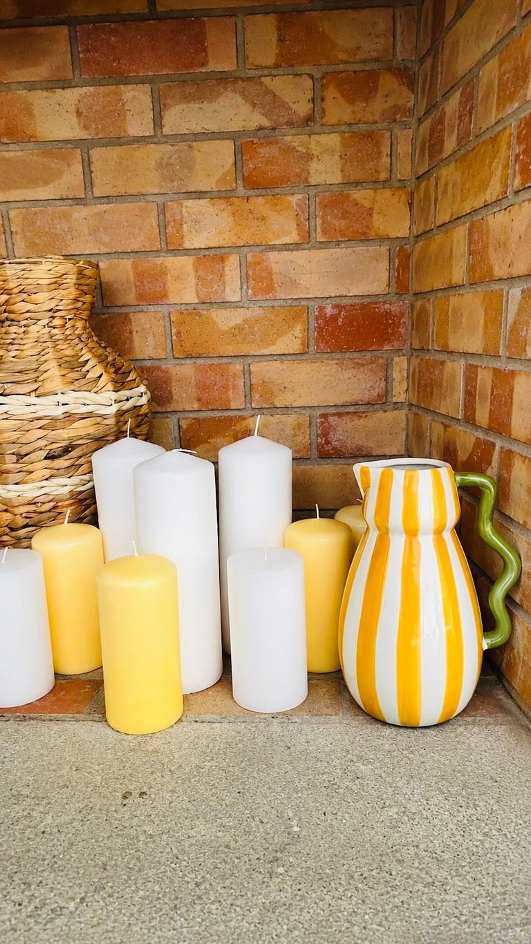 Fireplace hearth with yellow and white pillar candles, yellow striped pitcher, and woven basket against brick wall
