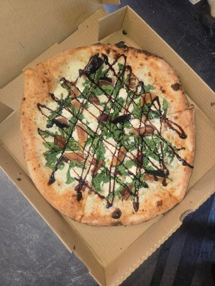 Gourmet pizza in cardboard box topped with fresh arugula, prosciutto, and dark drizzle sauce