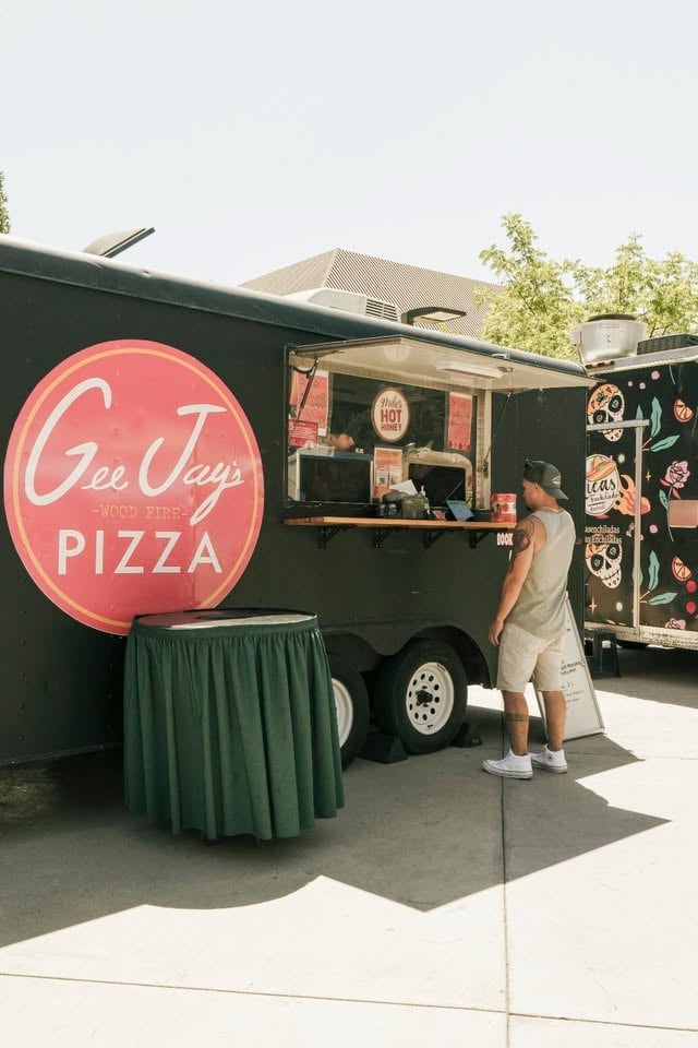 Green food truck with neon pink Gee Jays Pizza sign and customer at service window in sunny outdoor setting