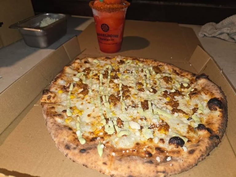 Artisanal pizza with white sauce, corn, and herbs in cardboard box with red beverage nearby