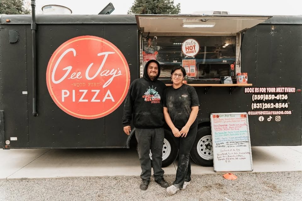 Two people standing in front of a dark green Gee Jays Pizza food truck with an illuminated neon sign and ordering information displayed.