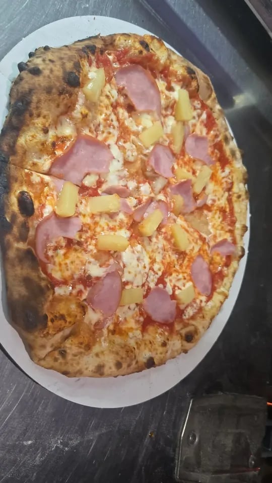 Wood-fired pizza with ham, pineapple, and cheese on a white plate and dark surface