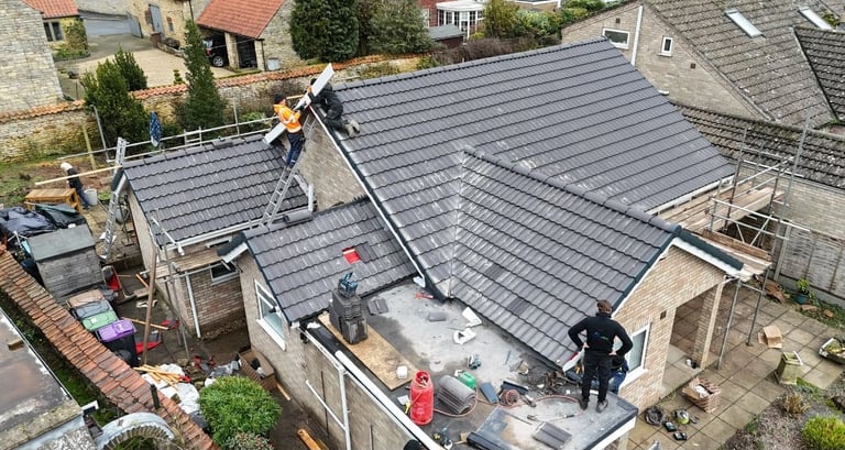 Aerial view of workers installing dark metal roofing panels on a residential house surrounded by neighboring properties