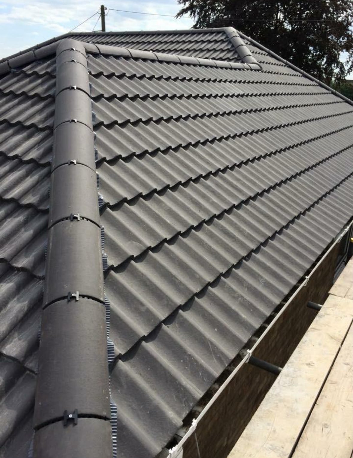 Angled view of a residential metal roof with gray corrugated tiles and a downspout, shot against a cloudy sky with trees in background