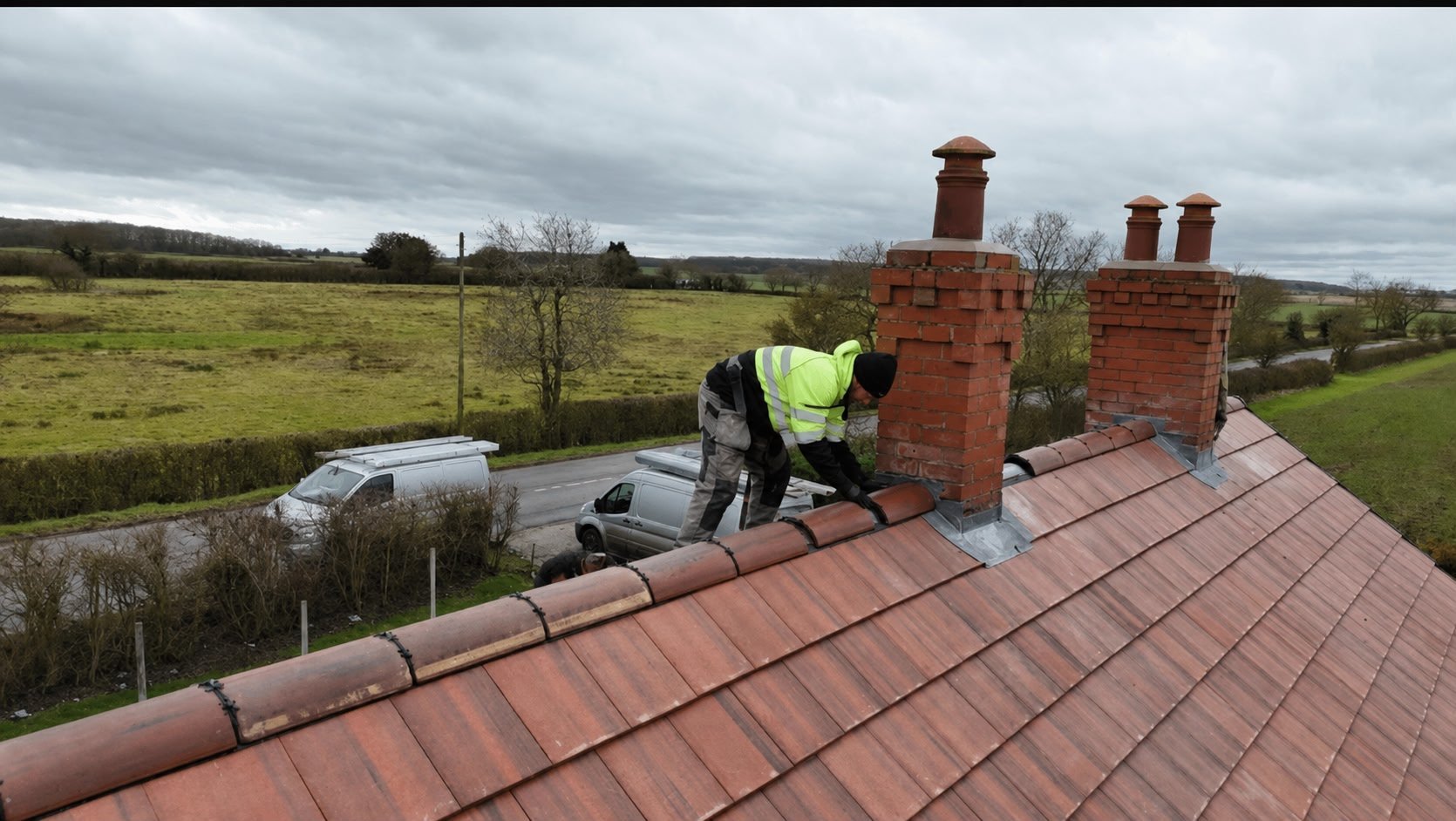 Worker in high-visibility vest on red tiled roof with brick chimneys overlooking rural countryside landscape