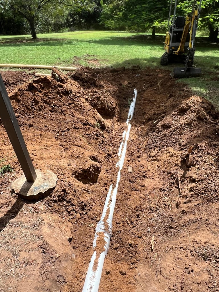 Excavation site showing a dug trench with white PVC pipe running through it, with yellow excavator in background on grassy area