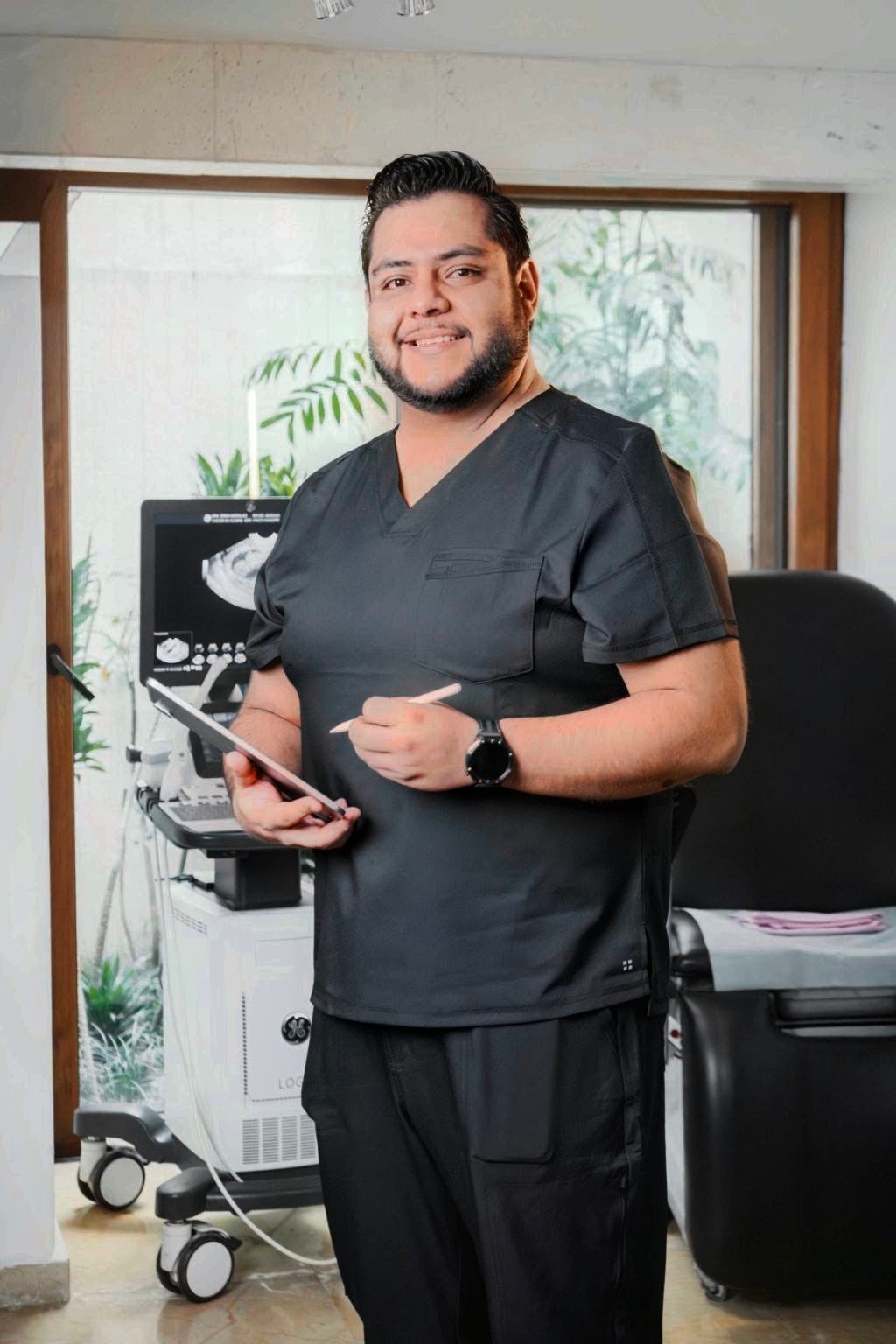 Profesional de la salud con uniforme negro sosteniendo una tablet en el consultorio, con equipo de ultrasonido al fondo