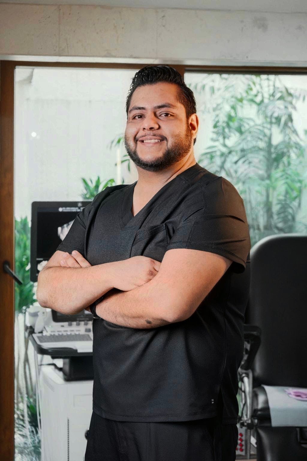 Hombre con uniforme médico negro y brazos cruzados sonriendo en el consultorio médico con plantas al fondo