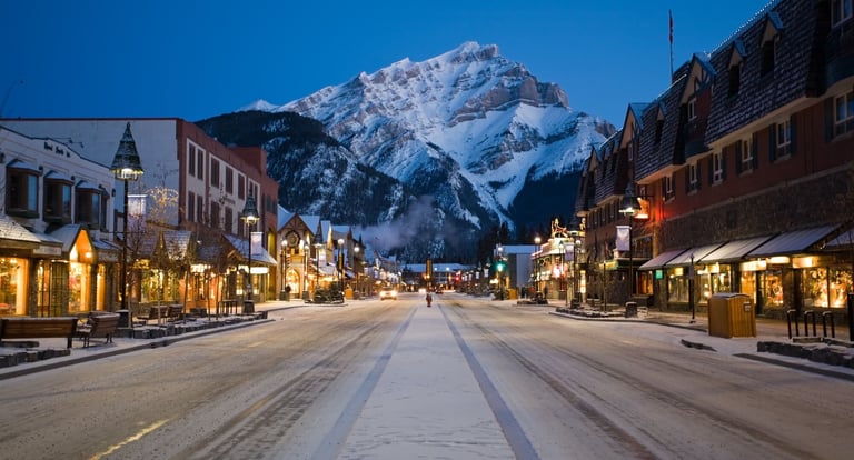Snow-capped mountain framed by a quaint Alpine town street at dusk with illuminated storefronts lining both sides